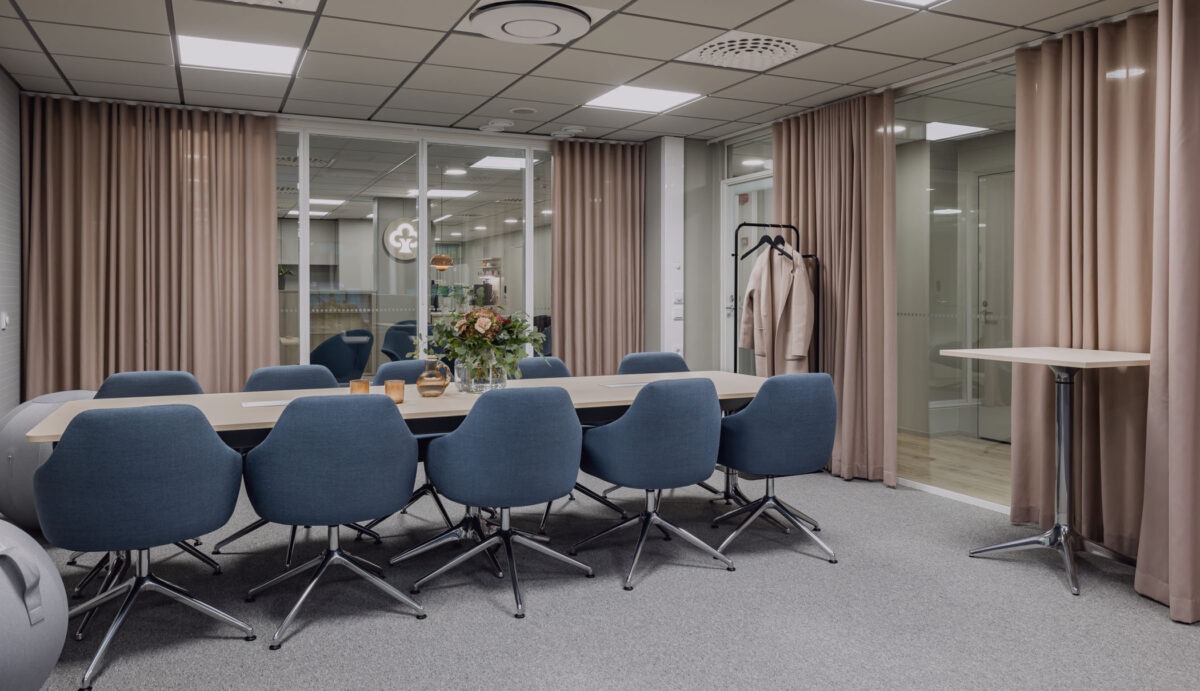 Kuvassa iso neuvotteluhuone, jossa iso pöytä, jonka ympärillä 10 sinistä tuolia. Huoneessa on lasiseinät, joita koristavat vaaleanpunaiset verhot. 